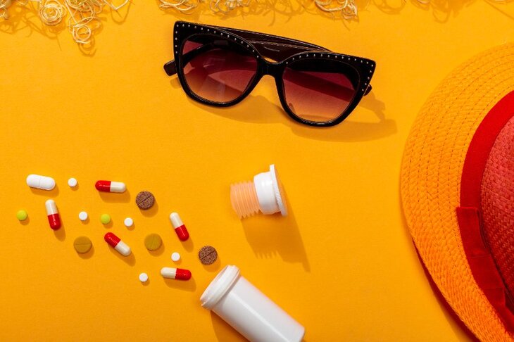 kompozycja na tle w kolorze żółcienia - okulary przeciwsłoneczne, kapelusz, tabletki rozsypane obok opakowania
