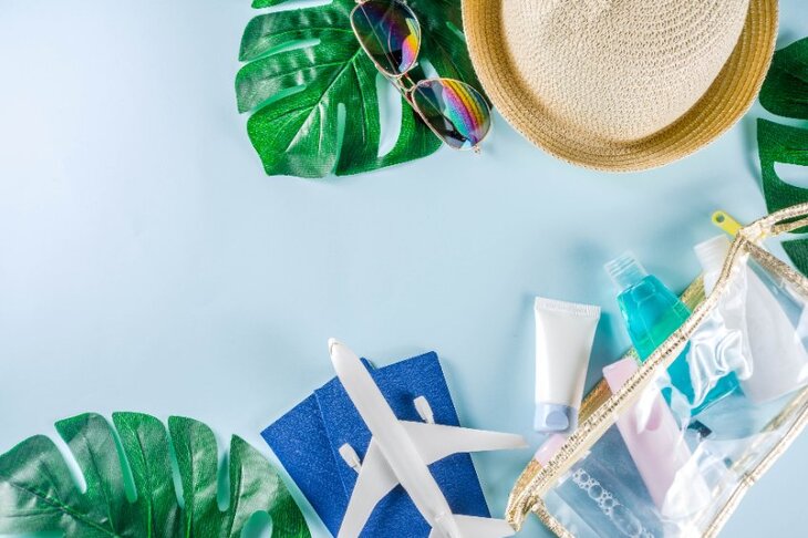 kompozycja na błękitnym tle - kapelusz, okulary przeciwsłoneczne, model samolotu, liście monstery oraz kosmetyki spakowane do samolotu do bagażu podręcznego