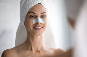Szczęśliwa piękna młoda kobieta z ręcznikiem owiniętym na głowie uśmiecha się do lustra. Na jej nosie znajduje się plaster oczyszczający.