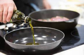 Zielonkawa oliwa jest nalewana na patelnię, w tle jest rozmyta druga patelnia, prawdobodobnie z mięsem na niej.