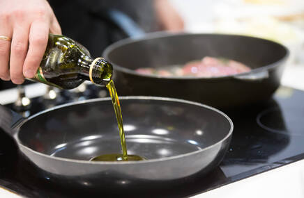 Zielonkawa oliwa jest nalewana na patelnię, w tle jest rozmyta druga patelnia, prawdobodobnie z mięsem na niej.