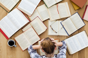 Kobieta siedzi przed biurkiem, na którym rozłożonych jest wiele książek, łapie się za głowę, bo czuje stres przed egzaminem