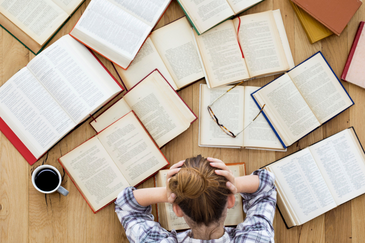 Kobieta siedzi przed biurkiem, na którym rozłożonych jest wiele książek, łapie się za głowę, bo czuje stres przed egzaminem