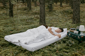 Mężczyzna śpi pod białą pościelą na materacu, który leży na mchu w lesie