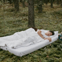 Mężczyzna śpi pod białą pościelą na materacu, który leży na mchu w lesie
