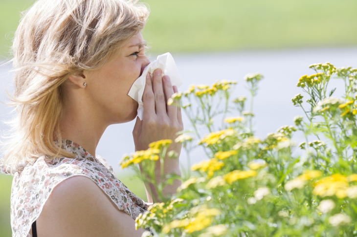 Kobieta wśród żółtych kwiatów wyciera nos chusteczką, bo ma alergię na pyłki