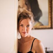 Młoda kobieta z jasnymi włosami upiętymi w luźny kok, ubrana w czarny top na ramiączkach, stoi przy białej ścianie.